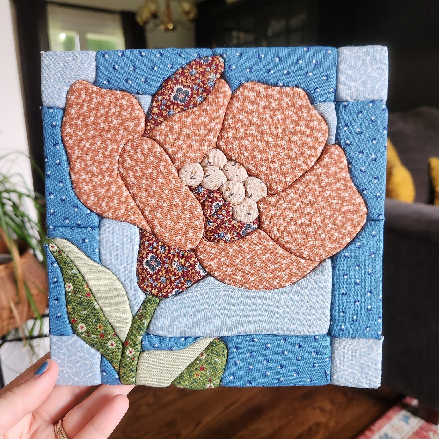 A handmade square quilted fabric wall hanging featuring a stylized flower with peach and light blue patterned petals, held up by a hand in a living room setting.