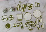 A flat lay of olive green and white die-cut craft embellishments, including bottle caps, a recycling symbol, a tree, leaves, and small decorative shapes, arranged on a white marble surface.