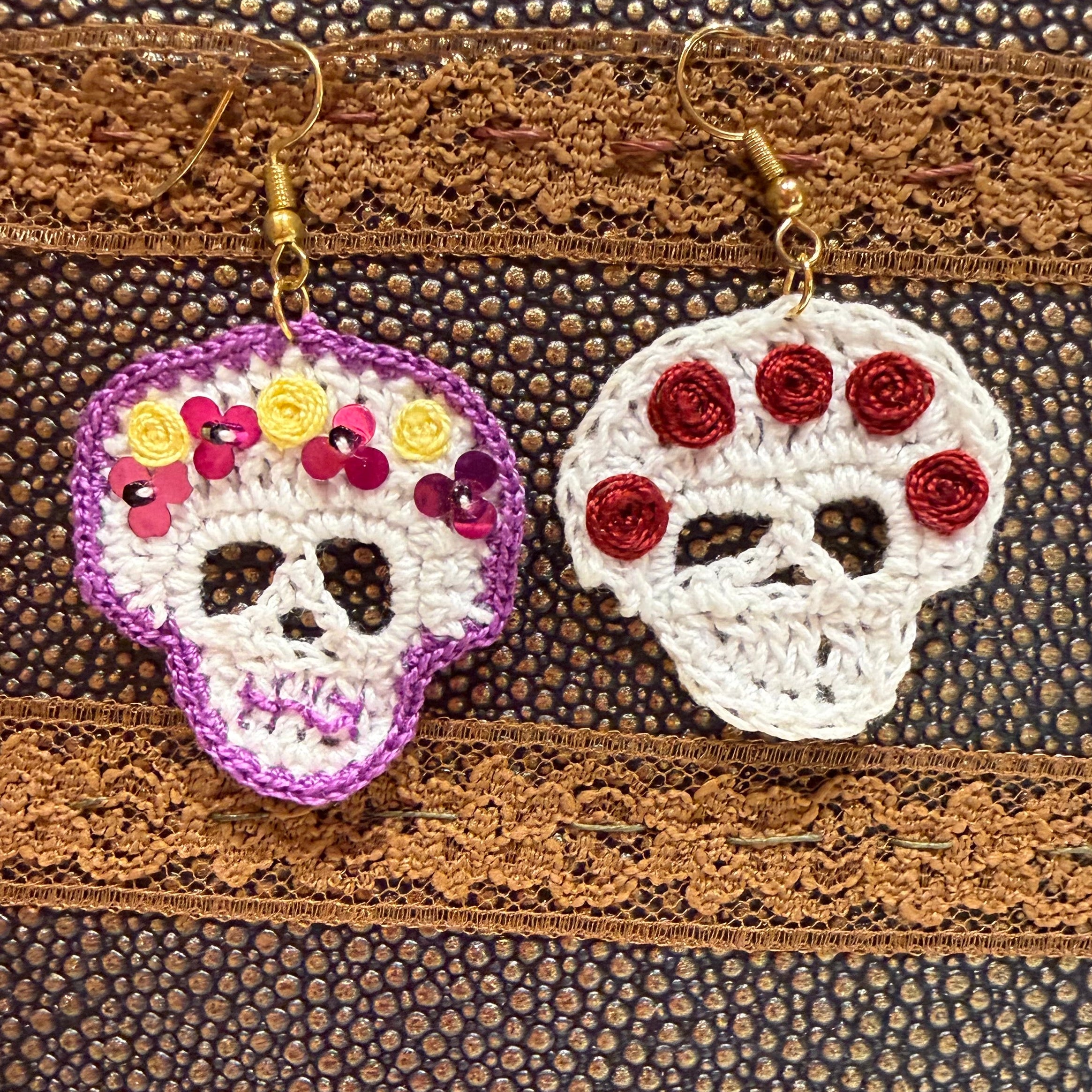 Two handmade crocheted sugar skull earrings hanging on a textured brown leather-like surface. One earring is white with purple trim and yellow, pink, and red floral details; the other is white with red rosette details.