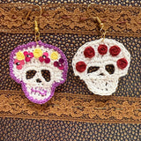 Two handmade crocheted sugar skull earrings hanging on a textured brown leather-like surface. One earring is white with purple trim and yellow, pink, and red floral details; the other is white with red rosette details.