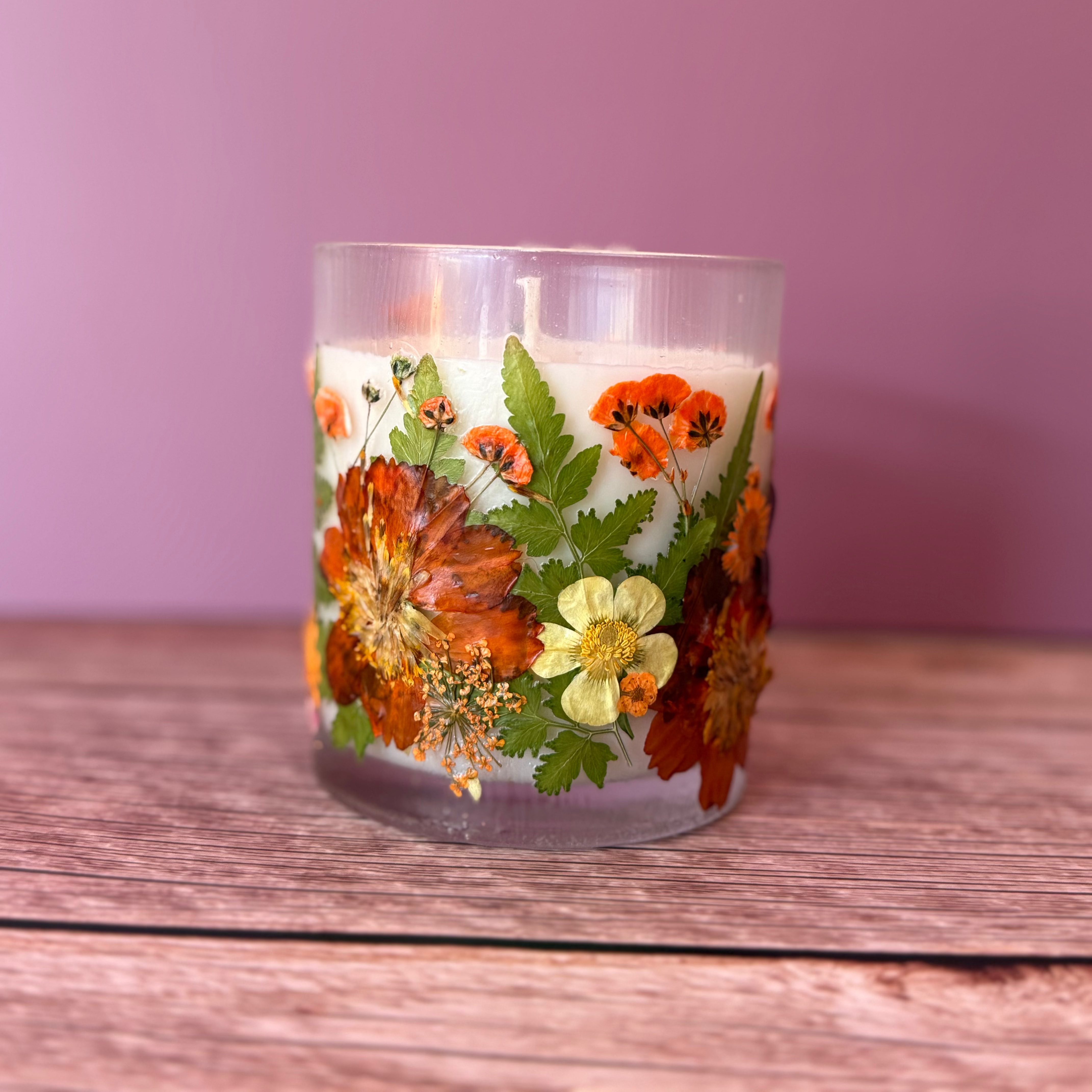 A clear glass jar candle filled with white wax and decorated with pressed orange flowers, green leaves, and small yellow blossoms, set on a wooden surface against a solid mauve-pink background.
