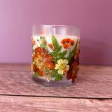A clear glass jar candle filled with white wax and decorated with pressed orange flowers, green leaves, and small yellow blossoms, set on a wooden surface against a solid mauve-pink background.