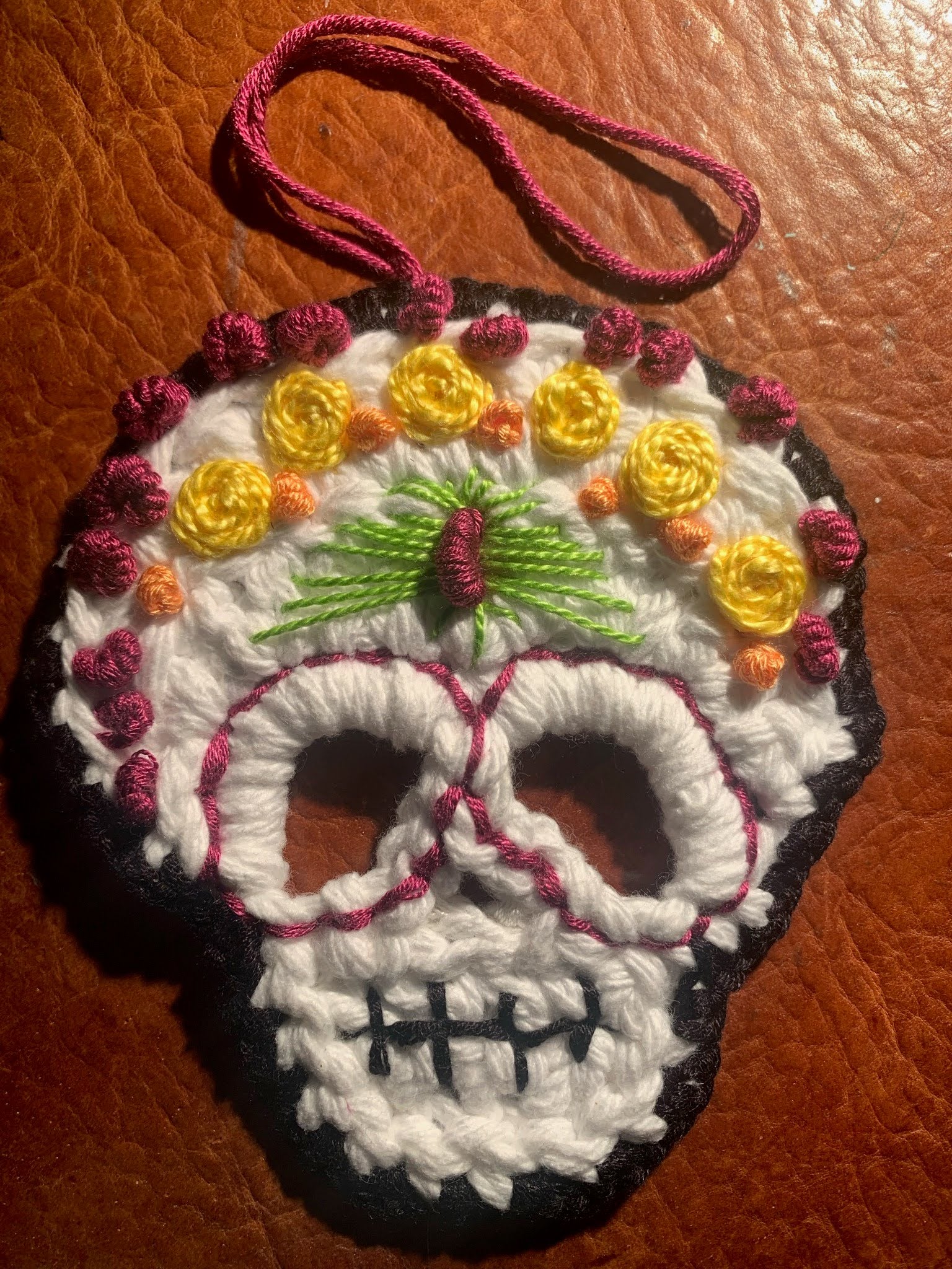 A handmade crocheted sugar skull ornament with a hanging loop, featuring white yarn for the skull face, yellow flowers, green leaves, and magenta and black accents, displayed on a brown textured leather background.